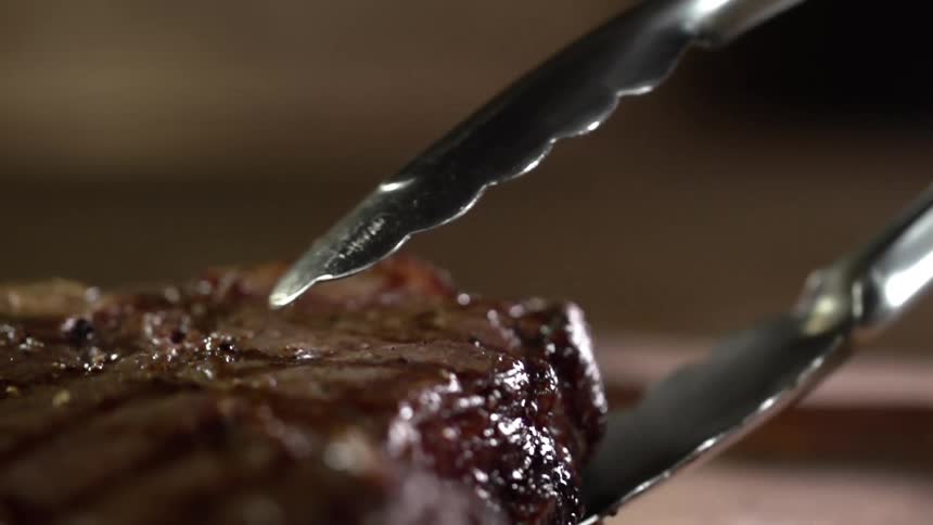 Close up of grilled steak being put on a steak board by a pair of tongs