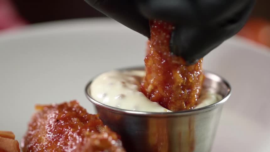 A gloved hand dips fried food into creamy sauce in a stainless cup on a plate