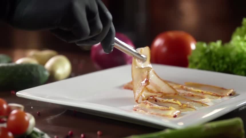 A person with gloves placing meat on a plate on a table surrounded by vegetables