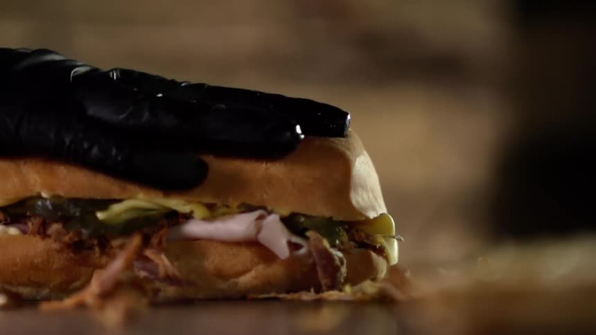 A person wearing a black disposable glove presses down of the top of a submarine sandwich