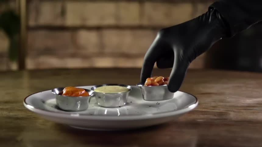 Person adds small metal bowl containing beans to others sitting on white plate on wooden table