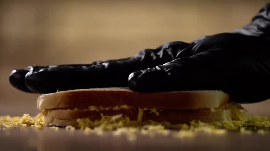 Hand wearing a black latex glove firmly presses down on two slices of white bread filled with shredded cheese and then releases the pressure