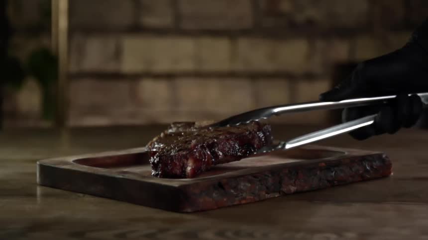 A person places a cooked steak on a rectangular wooden plate with a pair of tongs