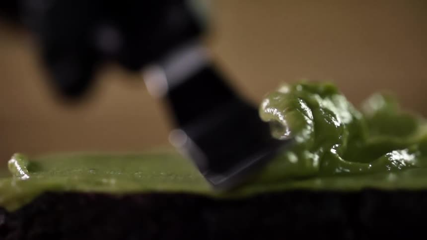 A gloved hand uses the back of a spoon to smooth an avocado puree on toast