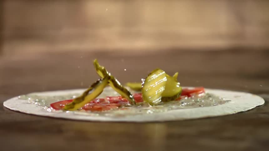 Pickle slices are being dropped on a wrap with sauces and tomato slices