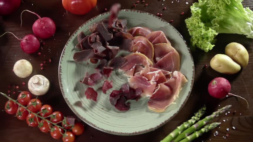 Food falling on to a plate with ham that is surrounded by tomatoes asparagus and potatoes