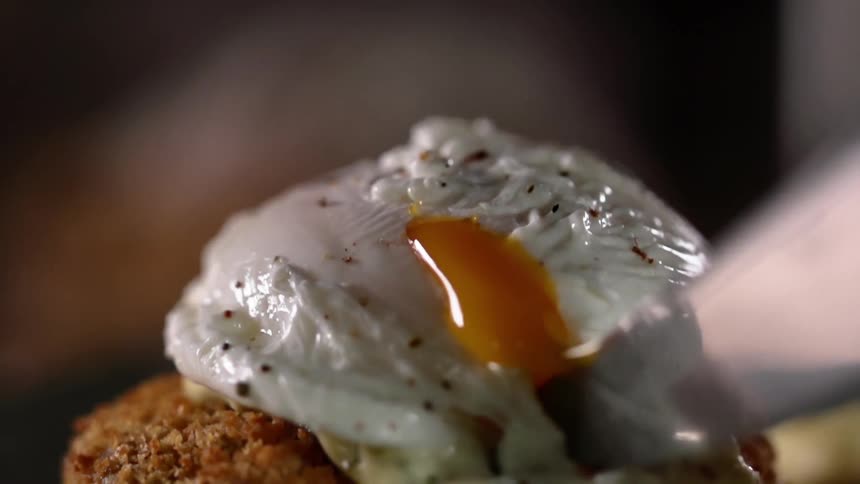 A knife cuts into a partially cooked egg sitting on top of a breaded patty