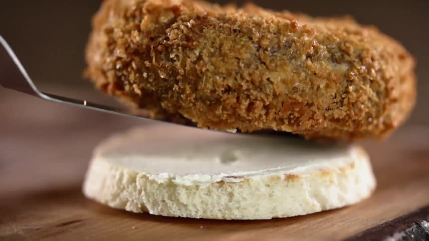 A metal spatula lowers a breaded patty onto a hamburger bun