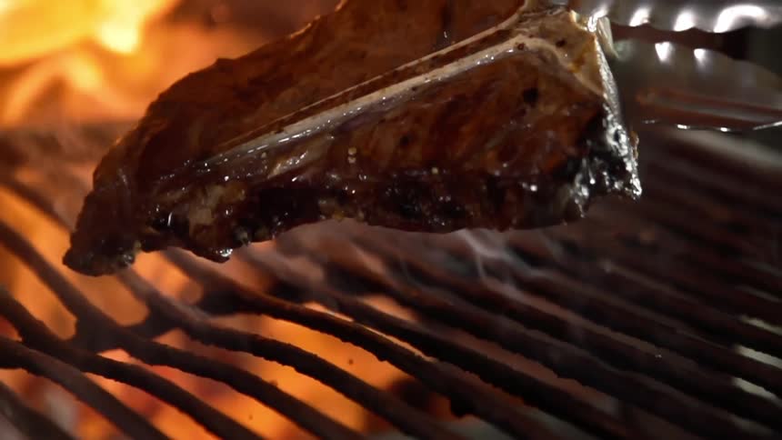 T bone steak grilling in the open flame is turned upside down with a metal tong