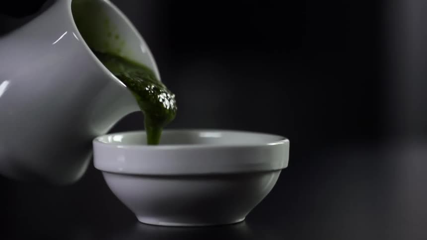 Green sauce pours from white ceramic pitcher into white ceramic bowl
