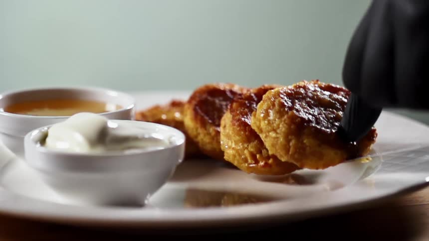 A gloved hand places crab cakes on a white plate next to two bowls of dipping sauce