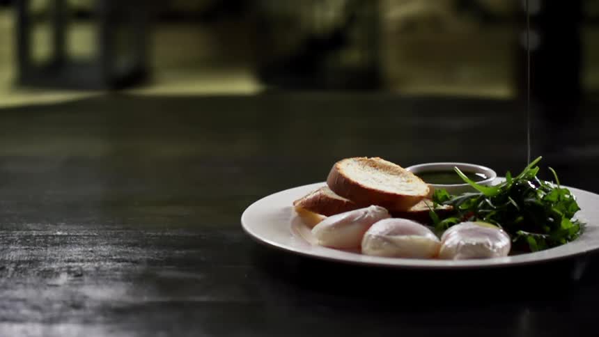 Dressing is poured onto greens on a plate next to poached eggs and bread