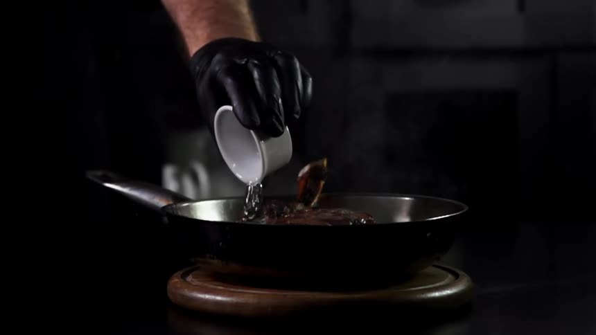 A person with gloves placing a liquid on a frying pan with meat inside of it with smoke coming off of it
