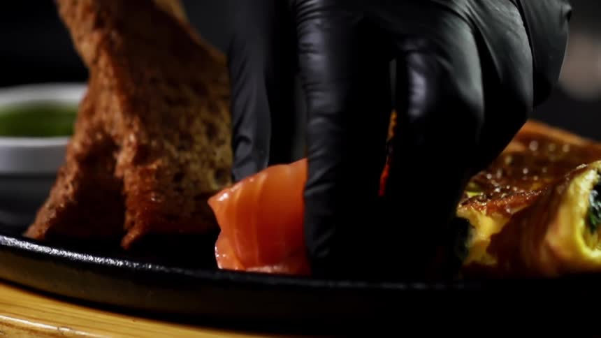 A person wearing black gloves places two pieces of raw salmon on a plate with bread and omelette