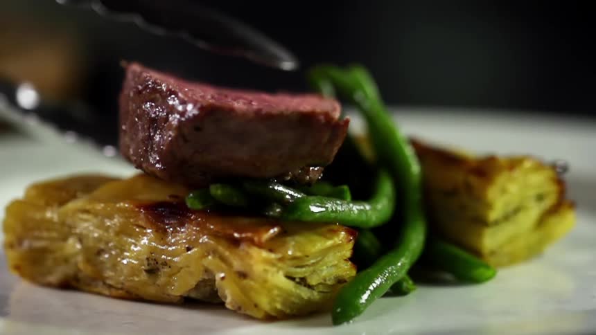 Tongs plate beef steak onto plate near green beans