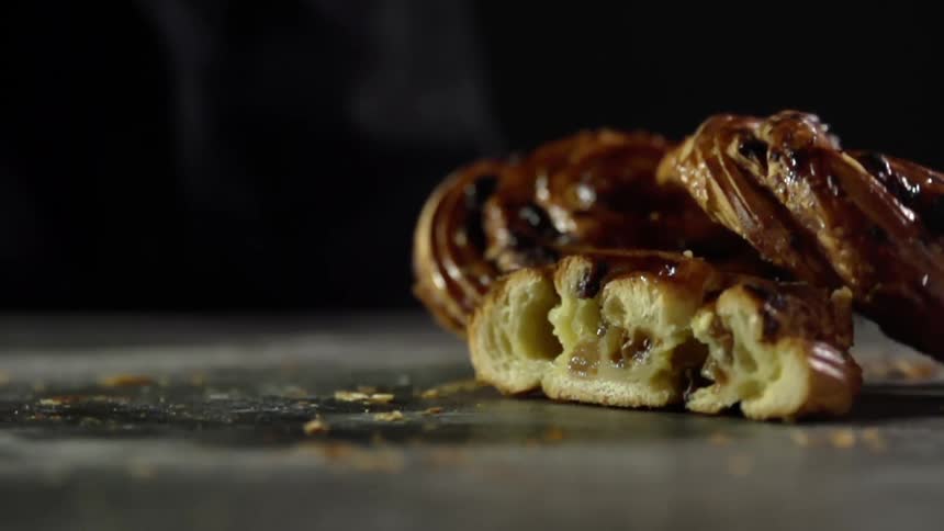 A glazed pastry falls atop similar pastries lying on a table covered in crumbs