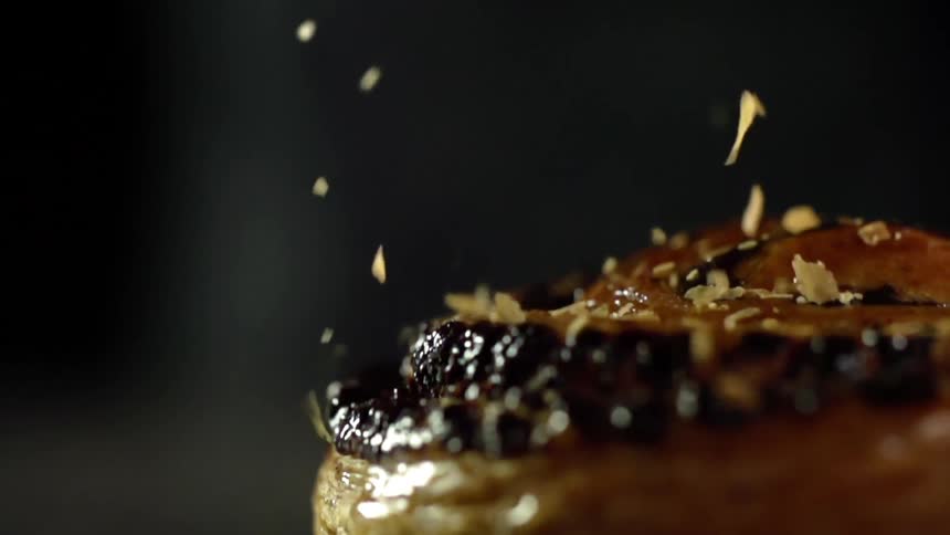 A bread pastry drops onto a surface in a spray of shaved nuts