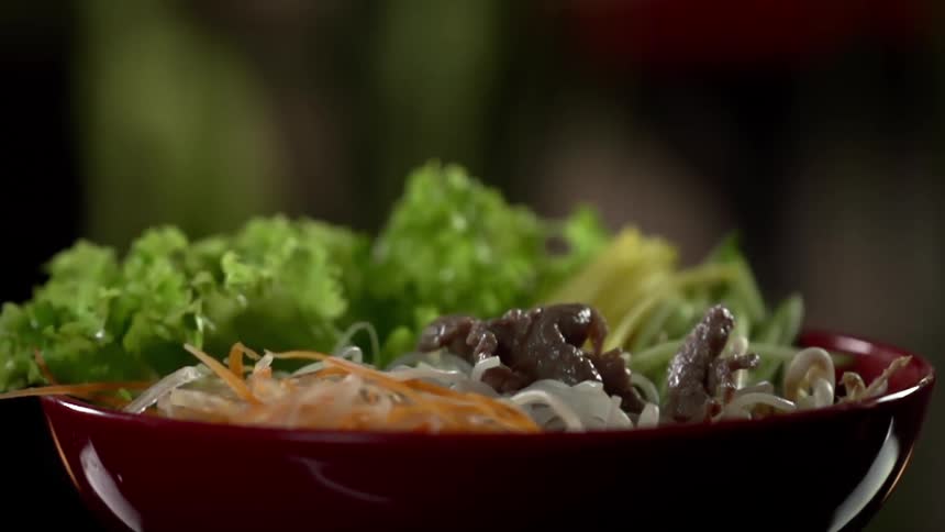 Cooked pieces of beef are dropped on a bowl with rice noodles and carrots
