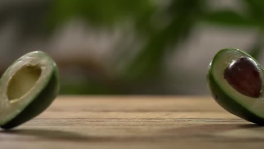 An avocado sliced in half drops on a wooden surface and falls apart