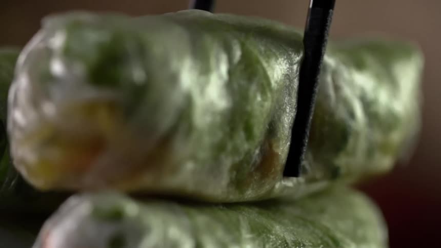 A pair of chopsticks are used to lift a spring roll off of a stack of other rolls