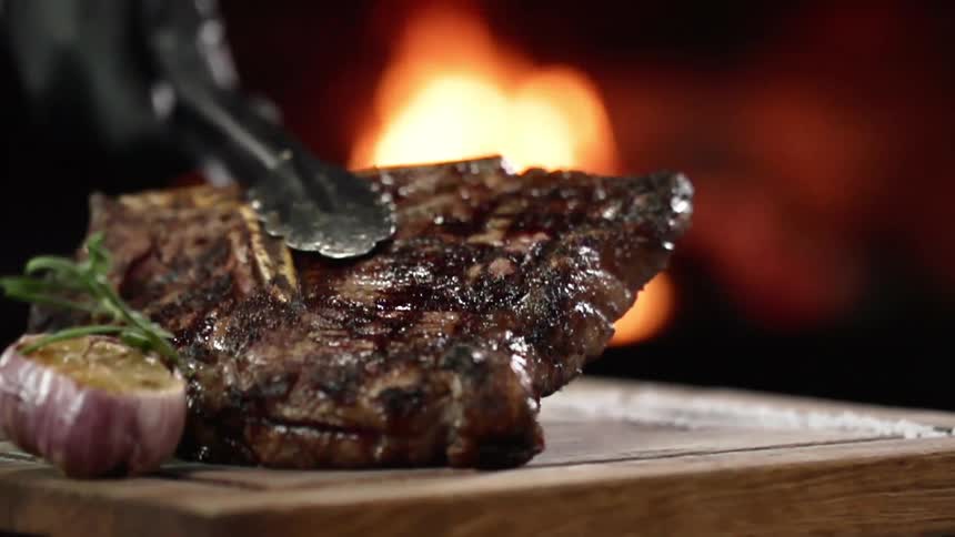 A cooked steak being placed on a wooden board with a pair of tongs as a fire burns in the background
