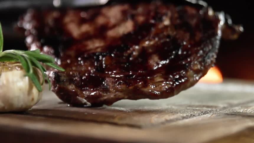 Metal tongs lower a cooked steak onto a wooden platter