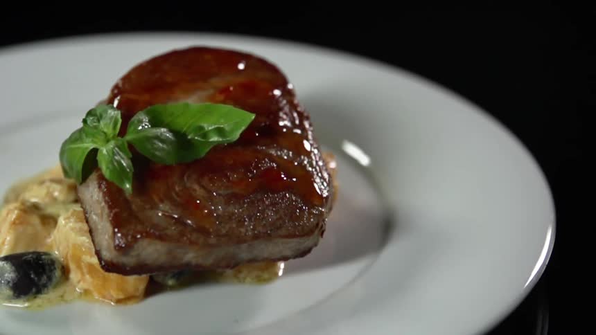 Green leaf falls over a glazed piece of cooked meat on a white ceramic plate