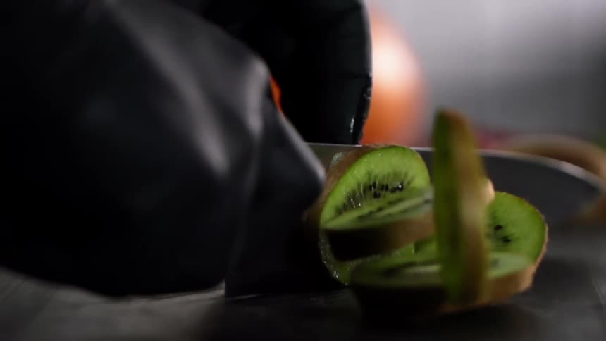 Hand wearing black latex gloves secures kiwi as another hand holding a chefs knife cuts the fruit into many slices