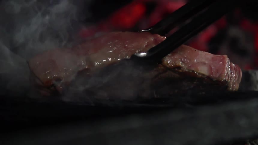 Steak cooking over a steaming hot grill picked up with tongs