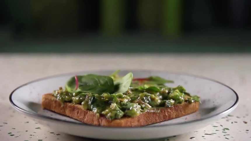 Several green leaves tumble down onto a piece of bread covered in spread on a plate