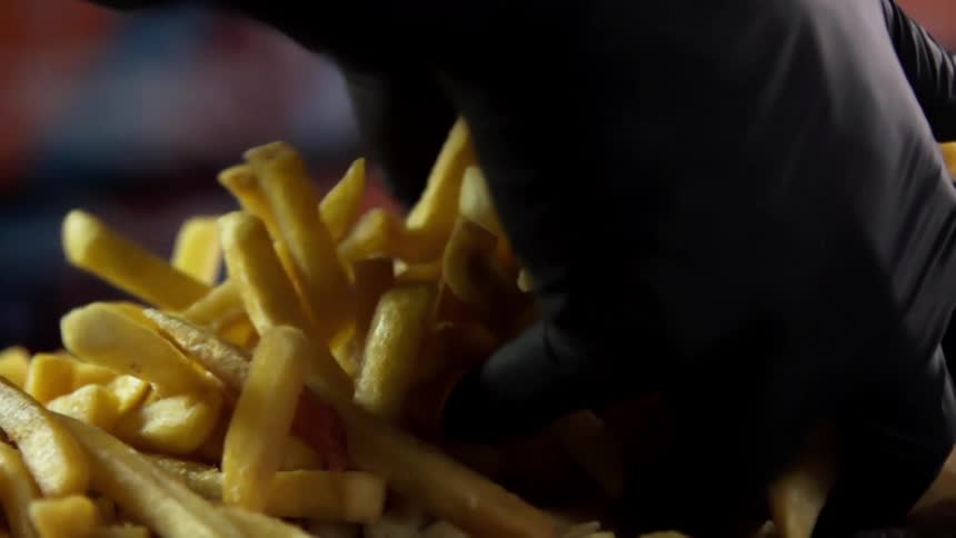 A paired of gloved hands reach into a pile of french fries and lift two handfuls of fries before letting them fall back onto the pile
