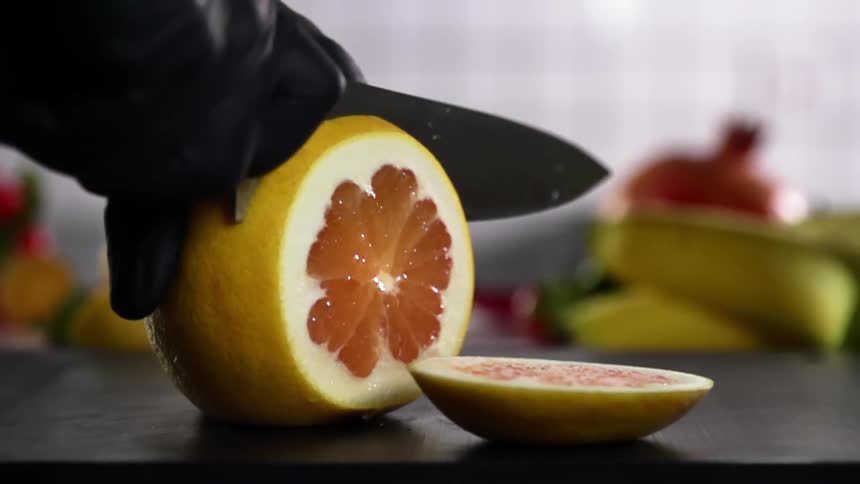 A person holds a piece of grapefruit and carefully slices it into pieces using a very sharp knife