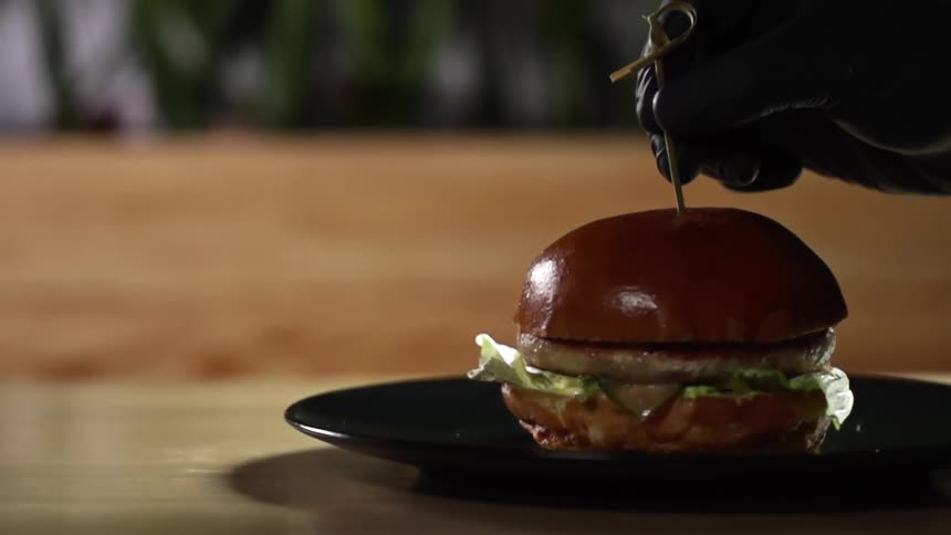 A person with a glove putting a skewer through a burger on a plate on a table