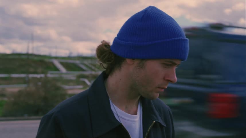 Man wearing blue knit hat stands while traffic passes on road between him and field containing large pipes and oil pump jack