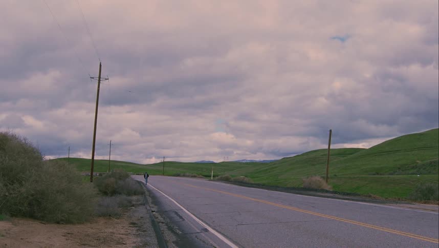 Person walks along shoulder of highway past shrubs and green hills under cloudy sky