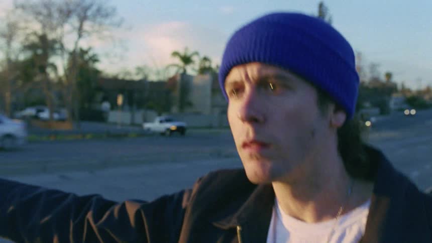 A young man in a jacket and blue knit beanie stares intently at the road while holding out his thumb in an attempt to hitch a ride