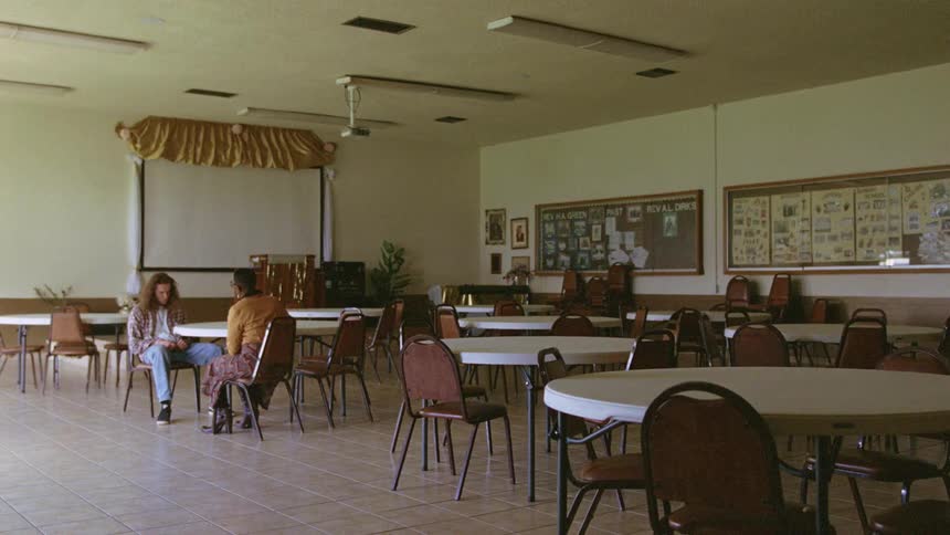 A man sits in a chair nervously tapping his foot while a woman sits across from him talking in an empty function room
