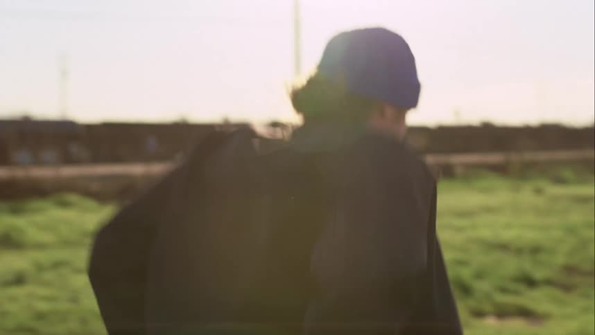 A man with a hat walking in a neighborhood with a grassy field with houses trees and the sky in the background