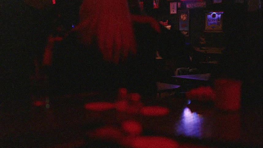 A man sitting on a chair next to a table in a bar with people standing in the foreground