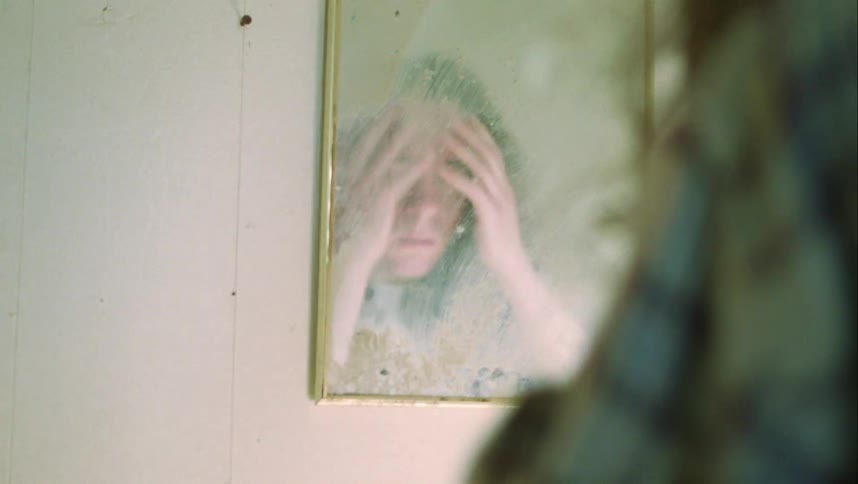 A teenager splashes water on his face and then looks in a dirty mirror and smoothes his hair back