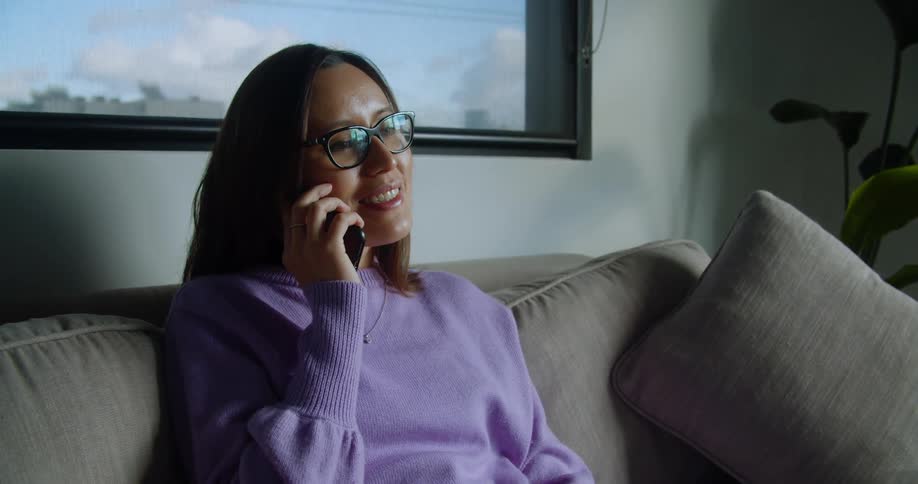A woman sitting on a gray sofa raises a smartphone to her face