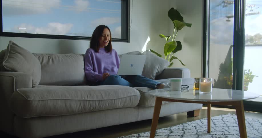 A woman sits on the couch talking as she uses her laptop