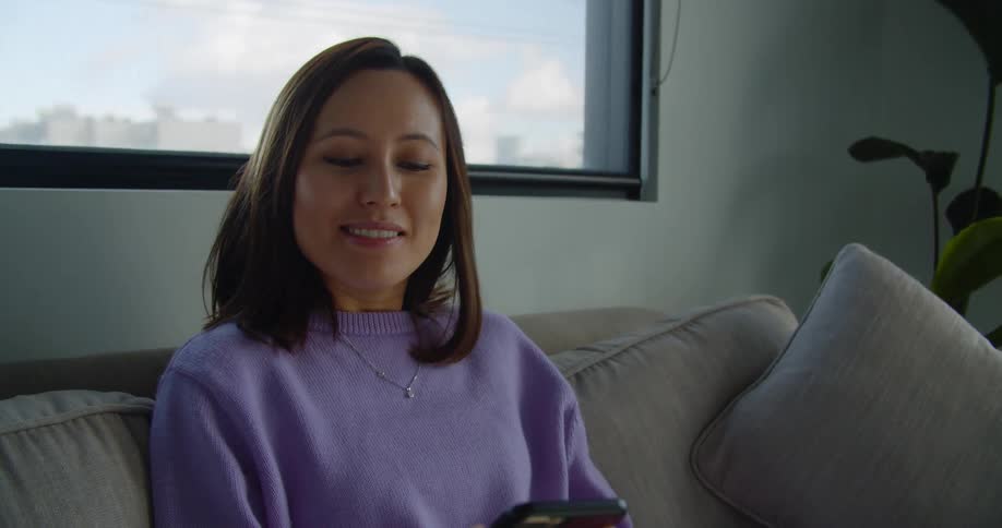 Woman wearing a purple sweater and jeans sitting on the sofa speaks on the phone and types on laptop in the living room
