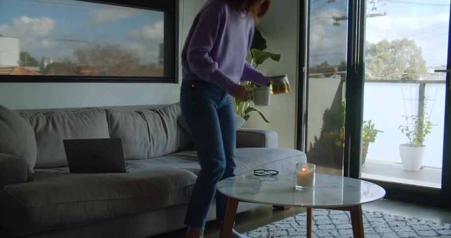 A woman walks toward a sofa and places a coffee mug and pot on a coffee table in front of it