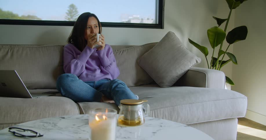 A woman sits at home on her couch with her laptop beside her and drinks tea