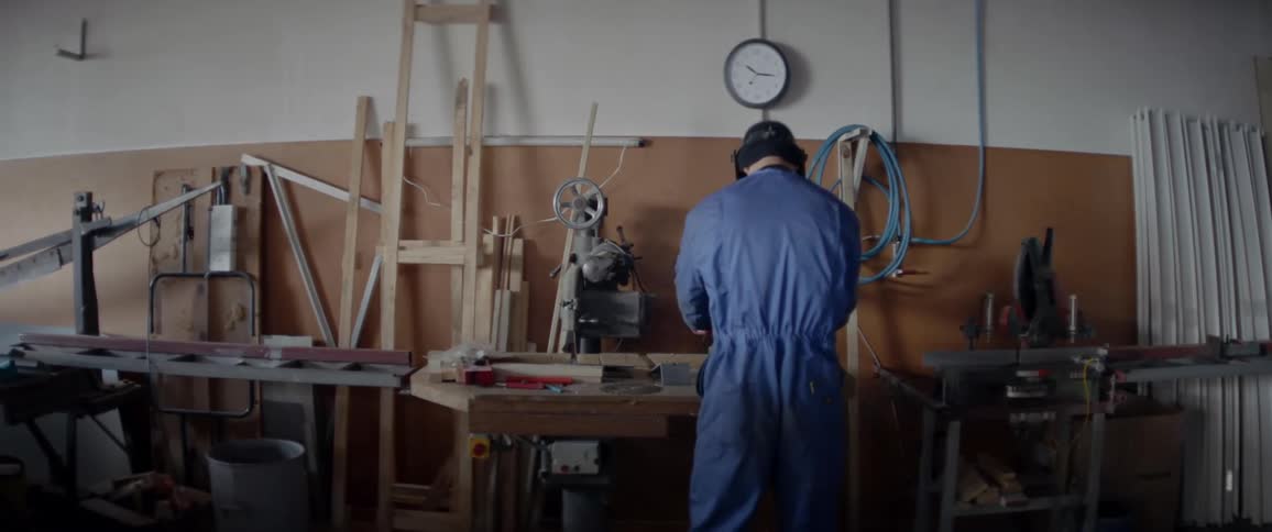 A man is flipping down his welding mask as he approaches a workbench during the day