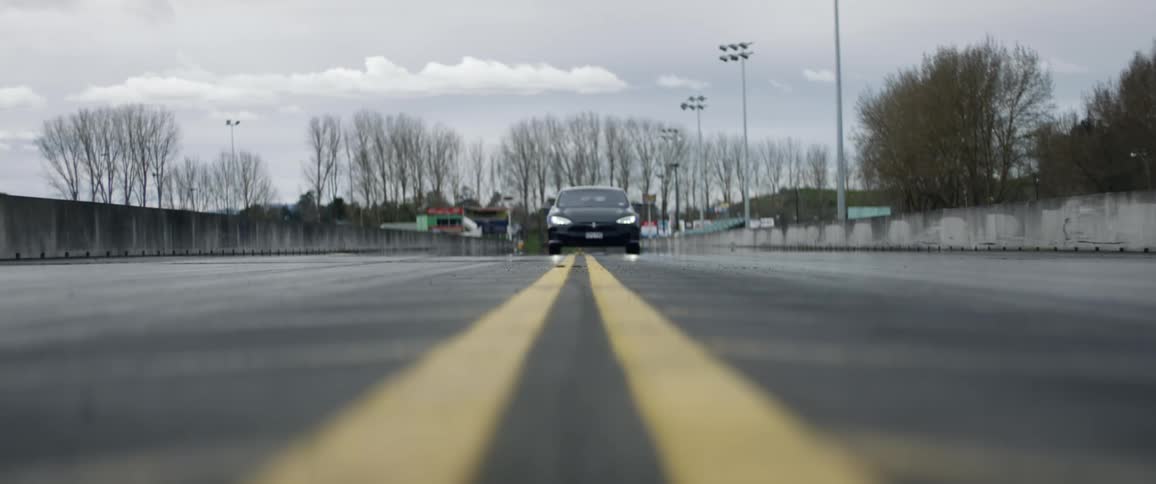 A car drives down the center of a road on a cloudy rainy day with bare trees in the background