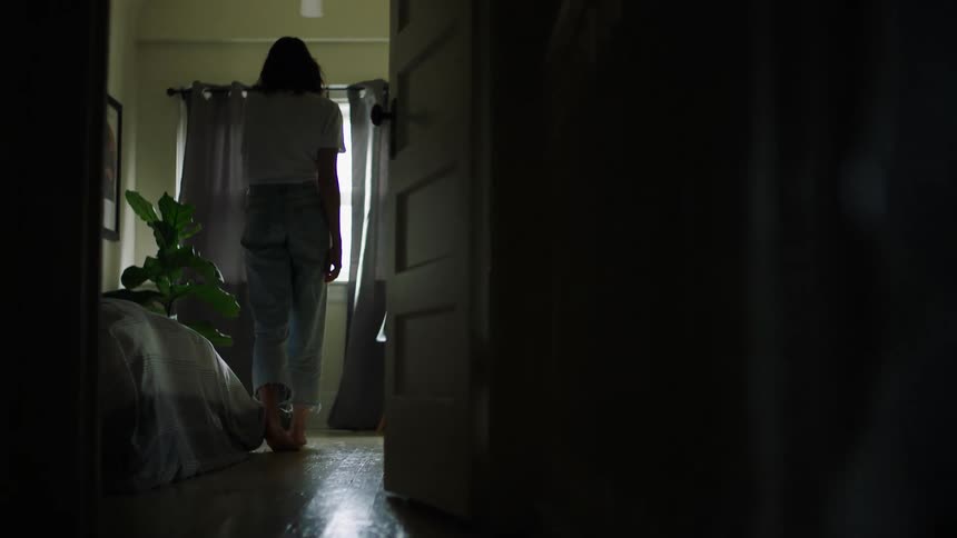 A woman rises from her seat on a bed a to approach a window and look out while lost in thought