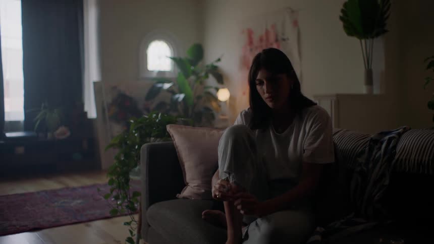 A woman sits on her couch in the dark, alone in a plant-filled loft.