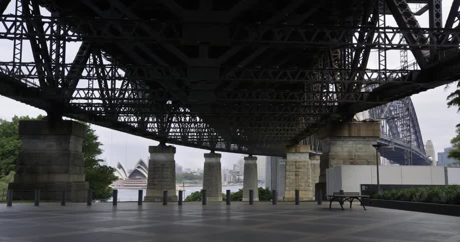 Underneath a bridge with a bench and pillars with a opera house and the sky in the background